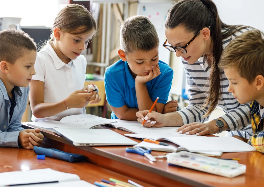 A female teacher helps four young students at a table in a classroom. They are all gathered around notebooks and papers, with the teacher pointing at the page with a pencil.